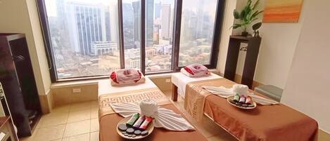 A room with two massage tables, each with towels and a tray of massage oils. Large windows offer a city view.