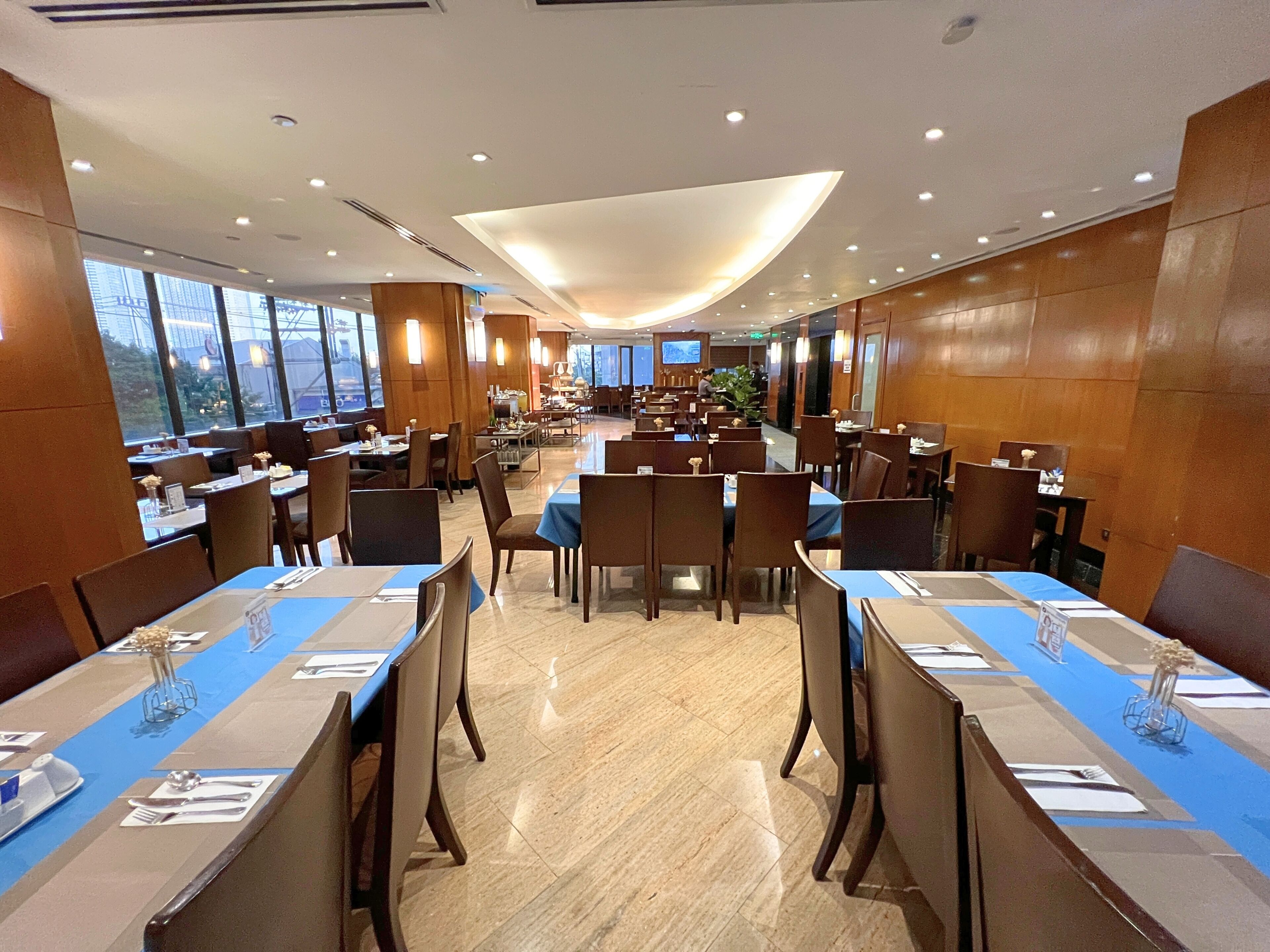 A spacious restaurant with multiple dining tables and chairs, large windows, and a modern ceiling design.