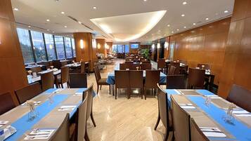 A spacious restaurant with multiple dining tables and chairs, large windows, and a modern ceiling design.