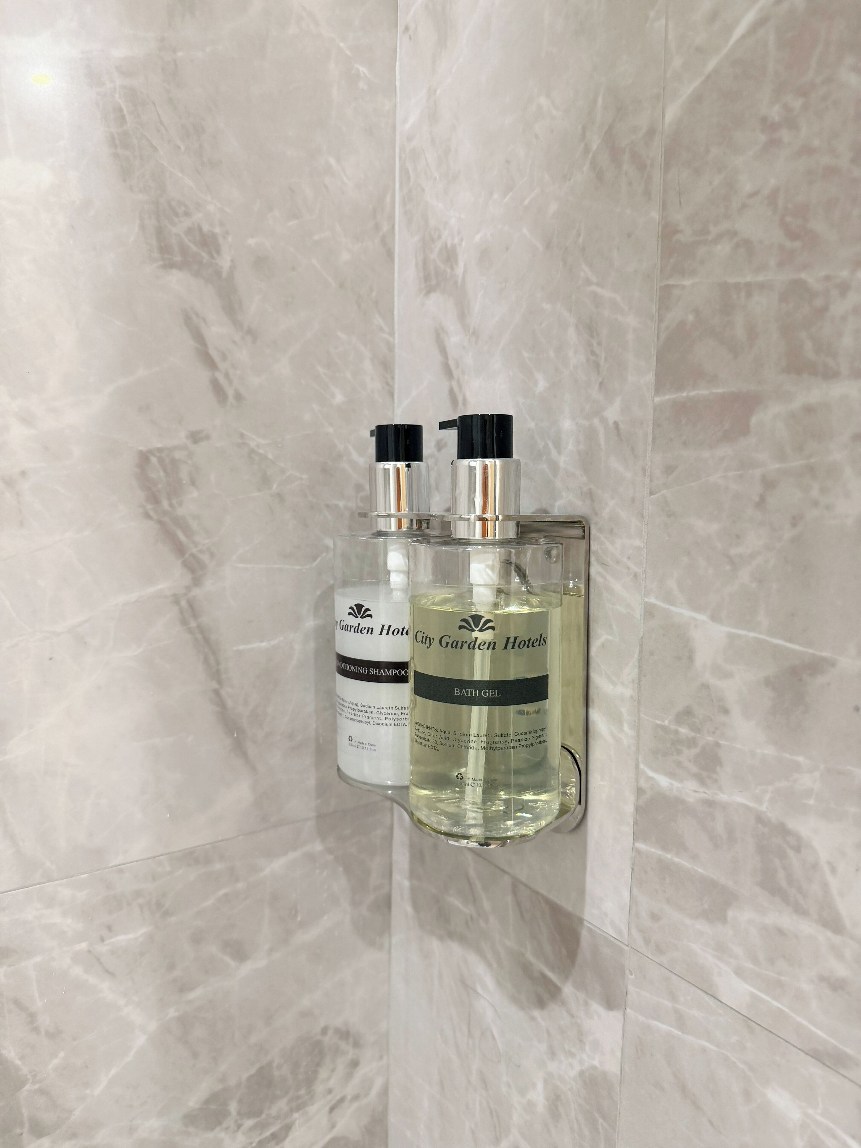 Deux flacons de gel douche et de shampoing après-shampoing de la marque City Garden Hotels, fixés sur un mur en marbre.