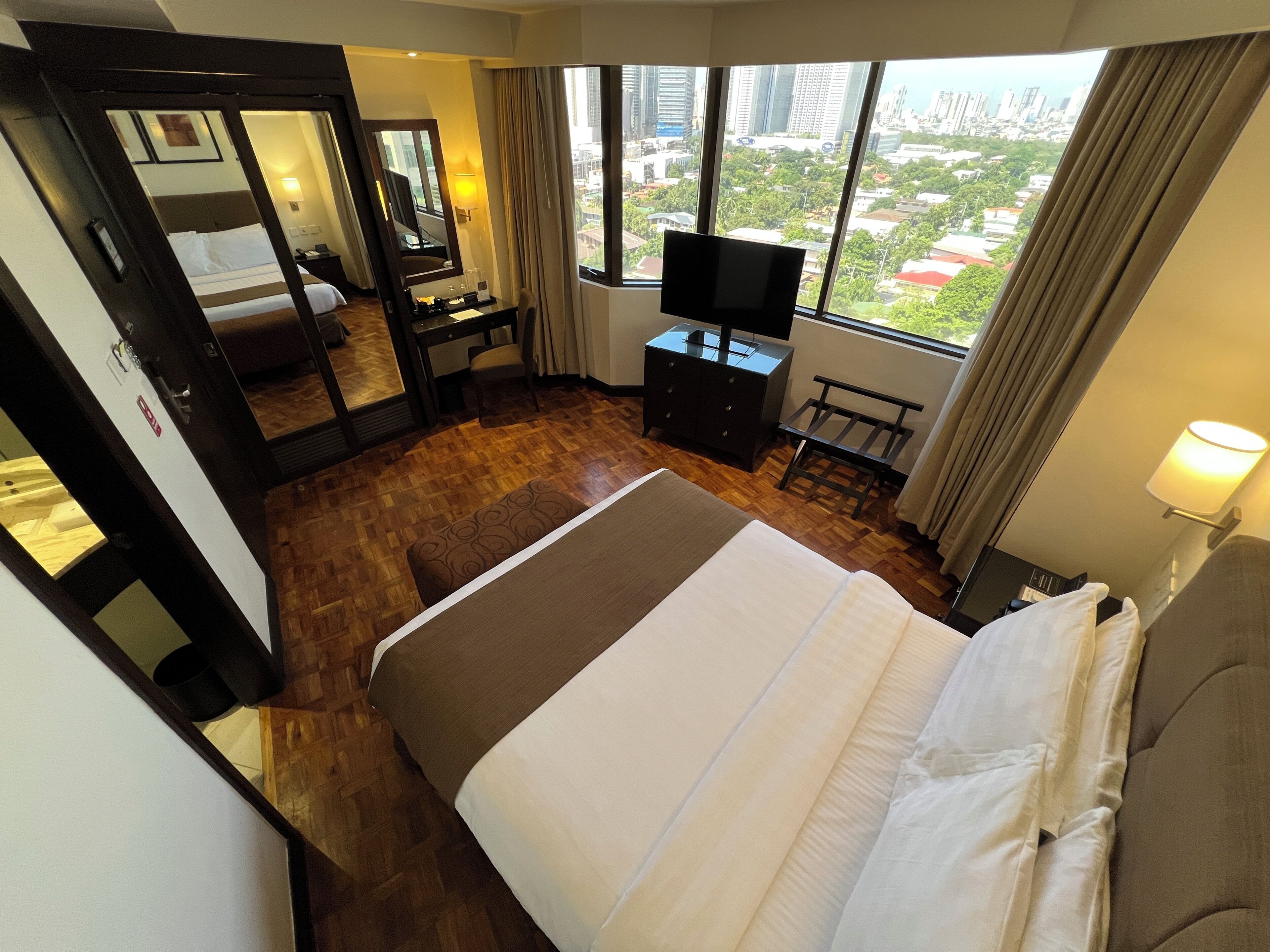 A hotel room with a large bed, a desk, a TV, and a view of the city.