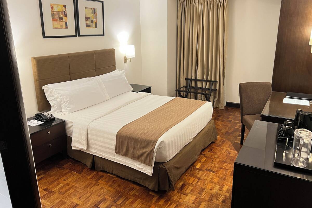 Habitación de hotel con cama, escritorio, silla, mesita de noche, lámpara y dos cuadros en la pared.