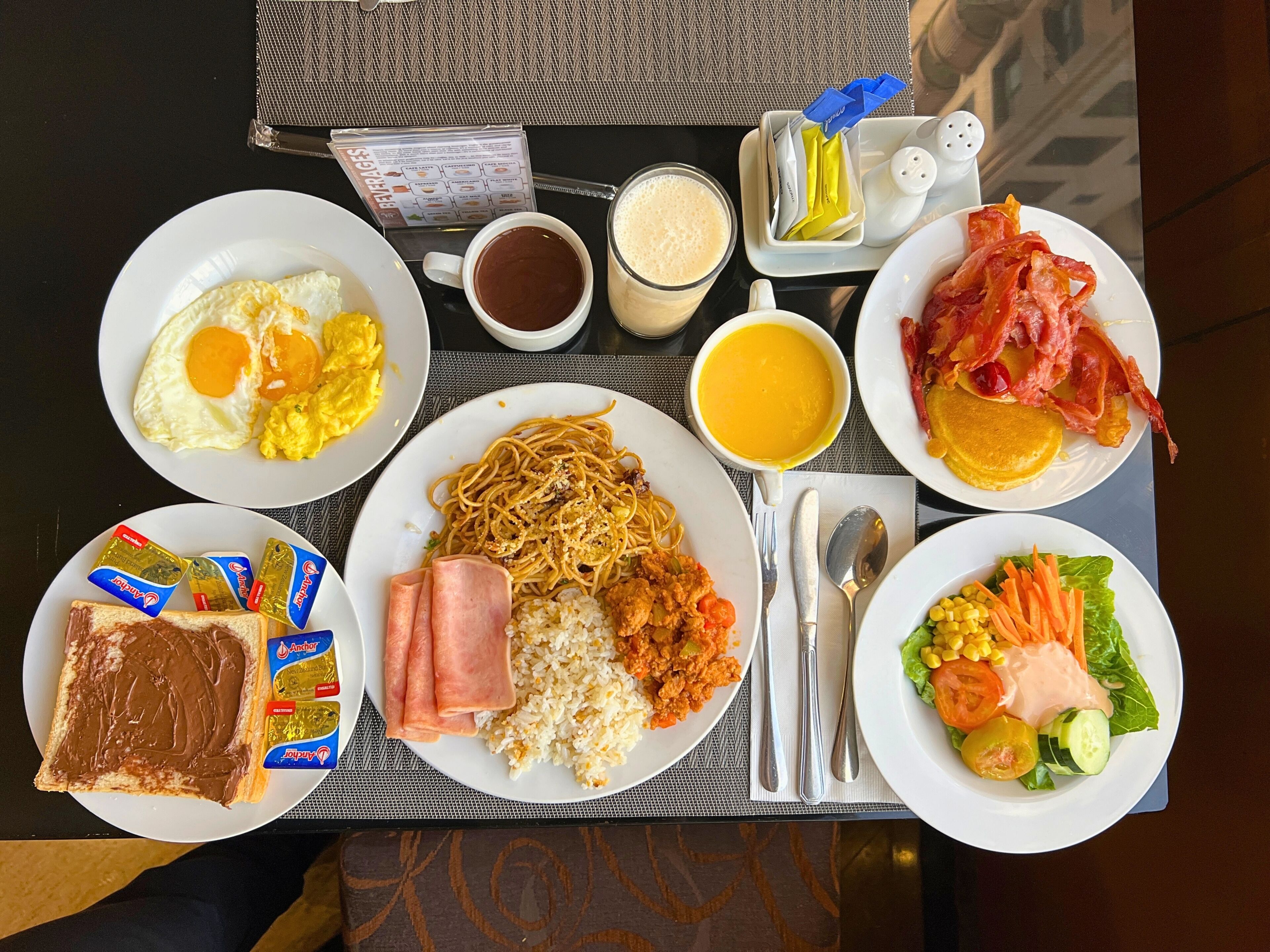 A table with breakfast items including eggs, noodles, rice, bacon, toast, and a salad.