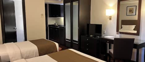 Habitación de hotel con dos camas, un escritorio con silla, un espejo, televisor y una kitchenette.