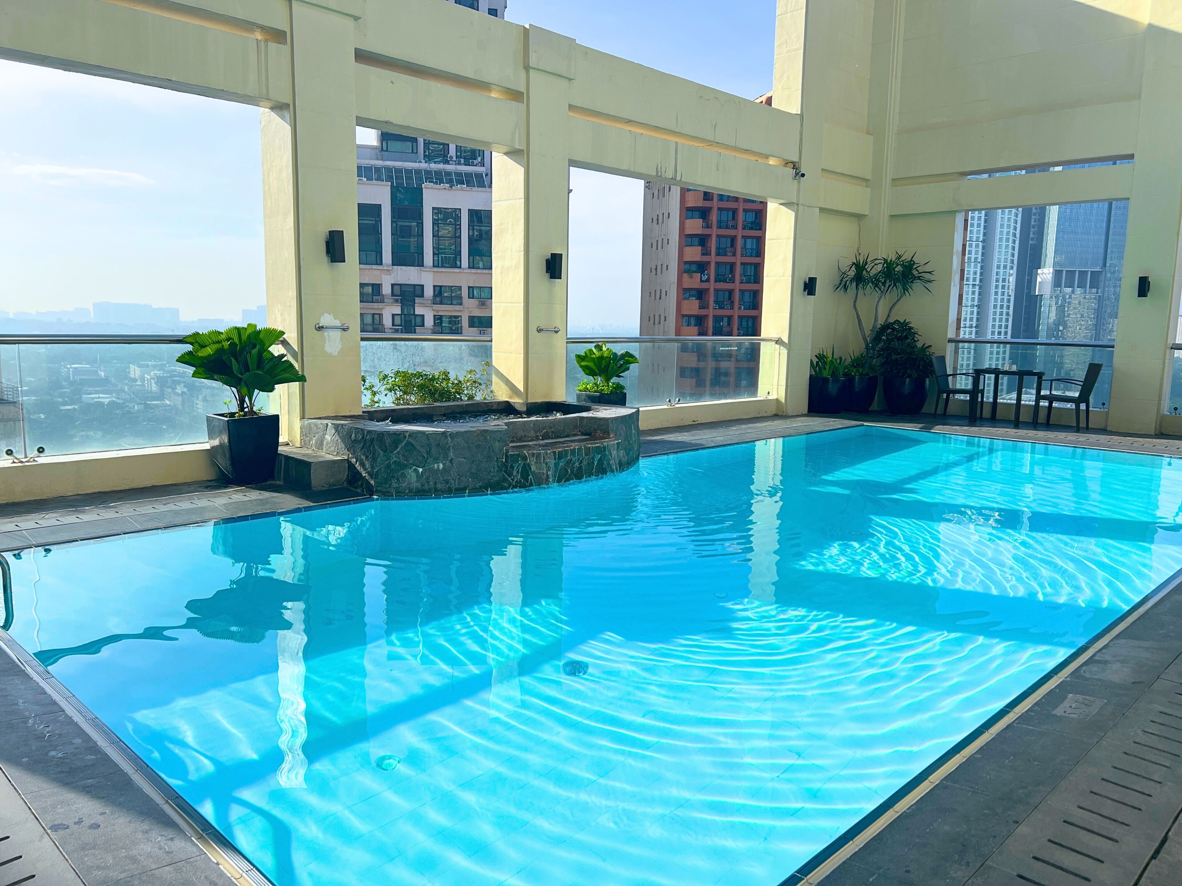 Una piscina en la azotea con agua azul cristalina, rodeada de plantas en macetas y una barandilla de vidrio.