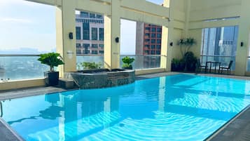 A rooftop swimming pool with clear blue water, surrounded by potted plants and a glass balustrade.