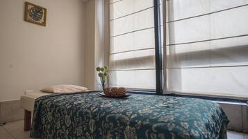 A bedroom with a bed, a window with blinds, a small plant, and a wall-mounted picture.