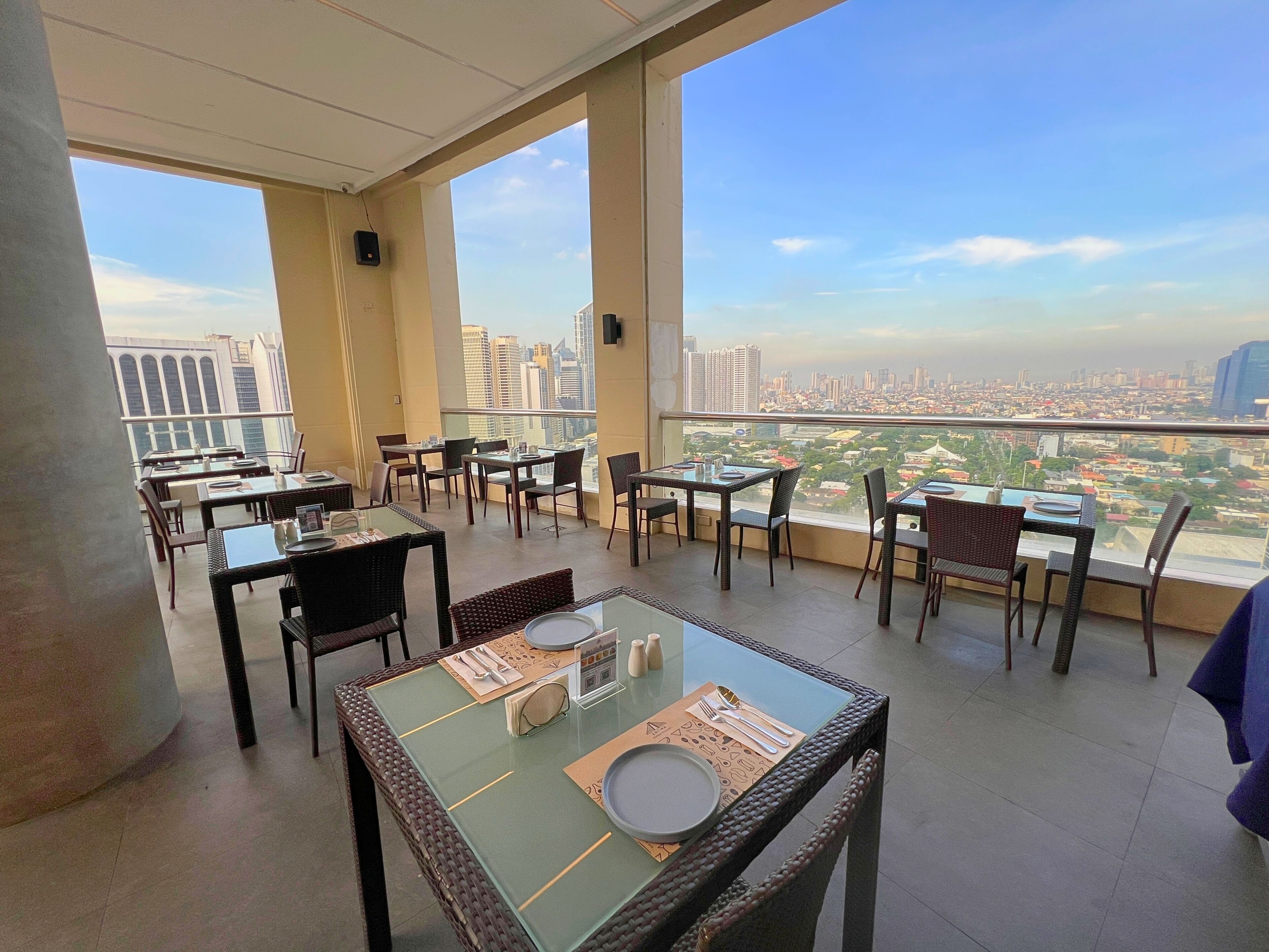 Un restaurante en la azotea con mesas y sillas, y grandes ventanales con vistas a la ciudad.