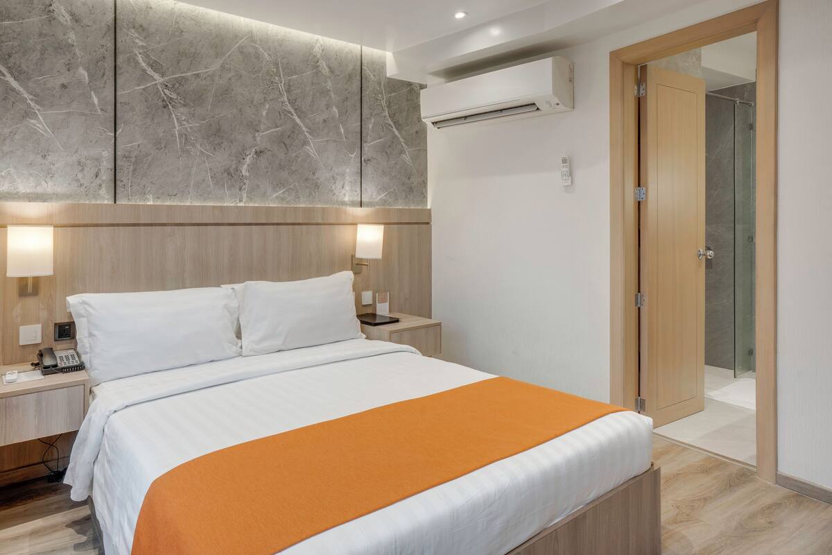 Habitación de hotel con cama, mesitas de noche, unidad de aire acondicionado, puerta de madera y ducha de vidrio.