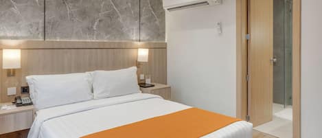 Habitación de hotel con cama, mesitas de noche, unidad de aire acondicionado, puerta de madera y ducha de vidrio.