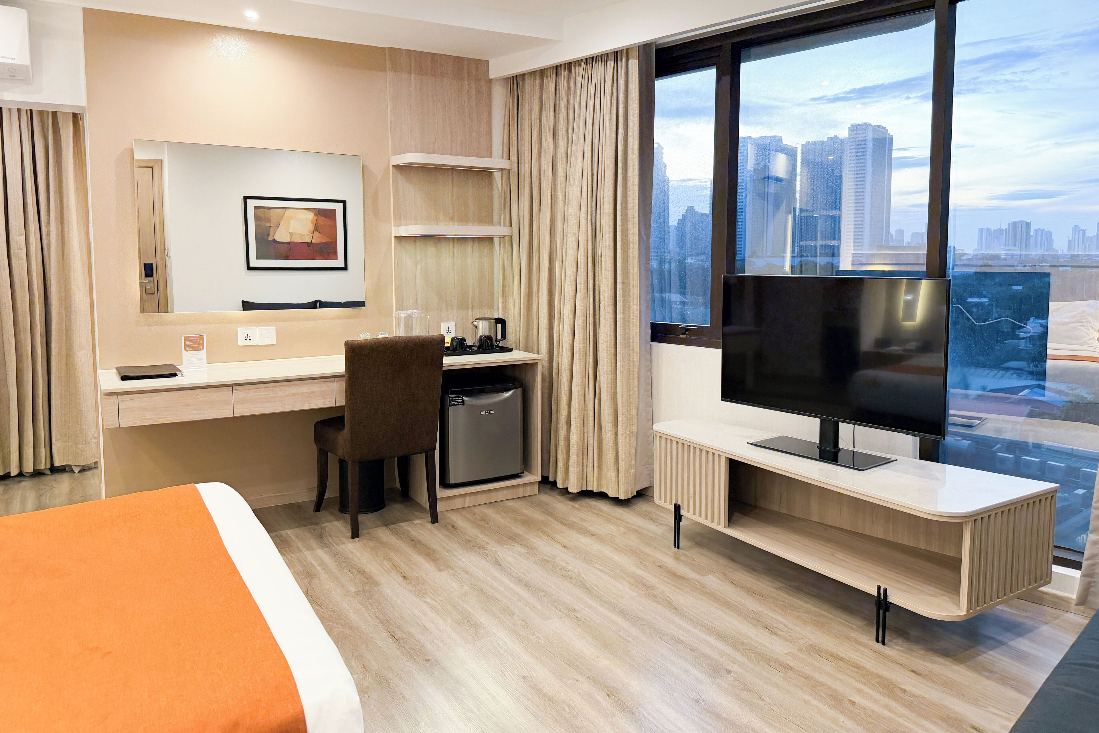 Ett modernt hotellrum med en säng, ett skrivbord, en stol, ett kylskåp, en TV och ett stort fönster med utsikt över staden.