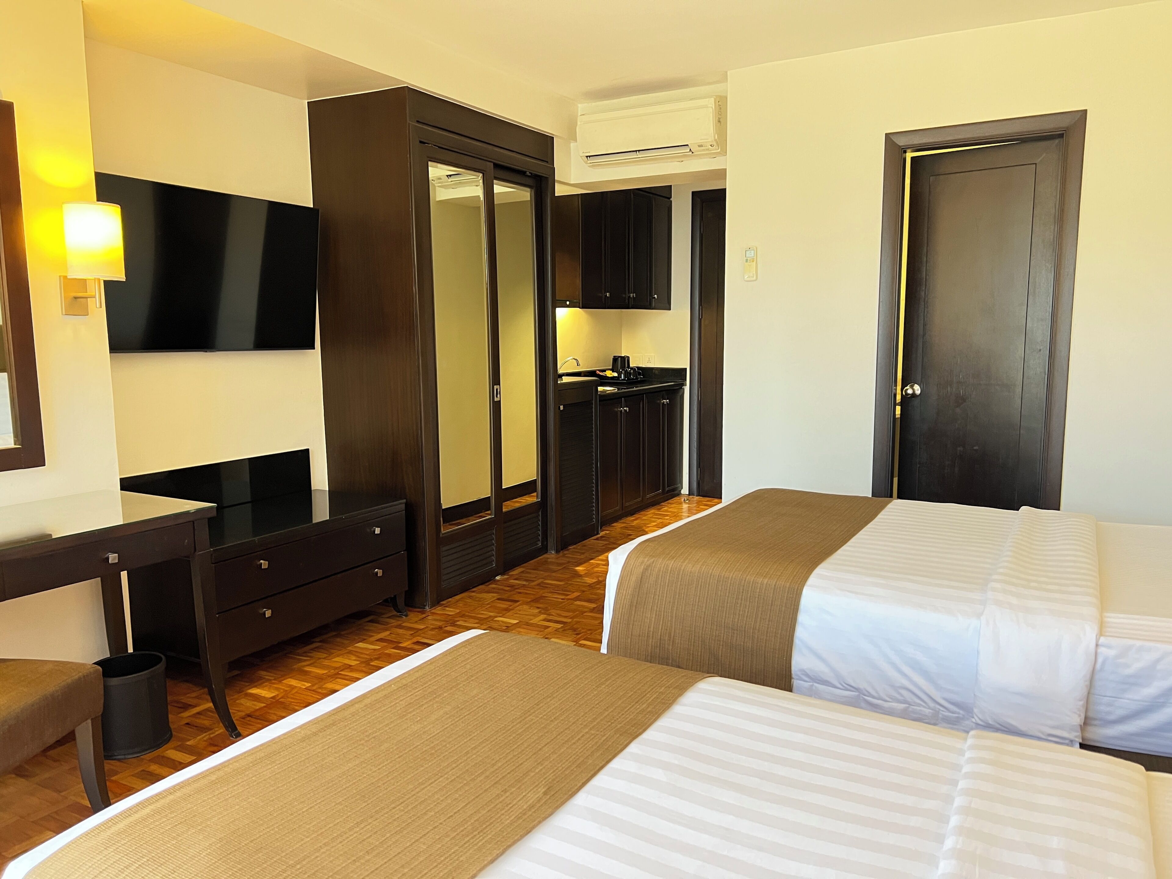 A hotel room with two beds, a desk, a mirror, a TV, a wardrobe, and an air conditioning unit.