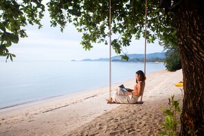 A person sitting on a swing under a tree by a beach with a view of the sea and distant hills.