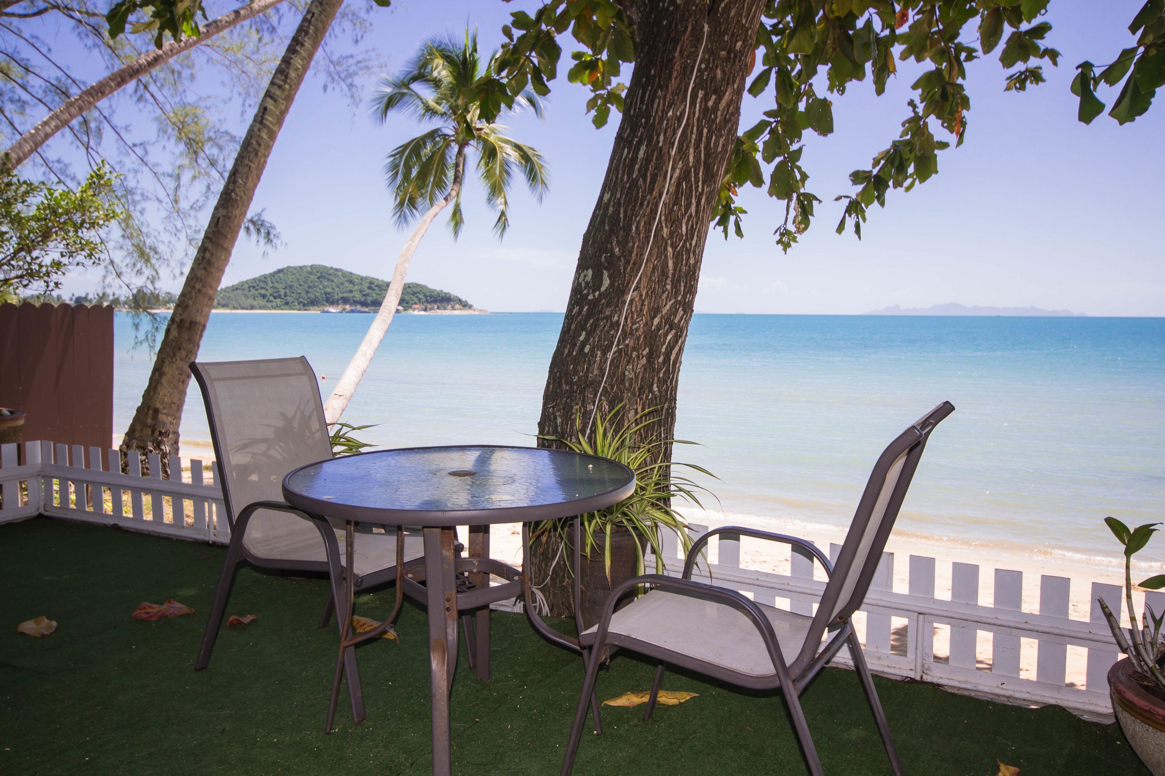 An outdoor seating area with a table and chairs, overlooking a beach with palm trees and a clear view of the ocean.