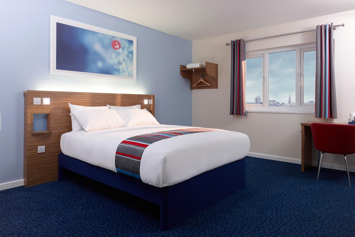 A hotel room with a bed, a desk, a chair, a window with curtains, and a painting on the wall.