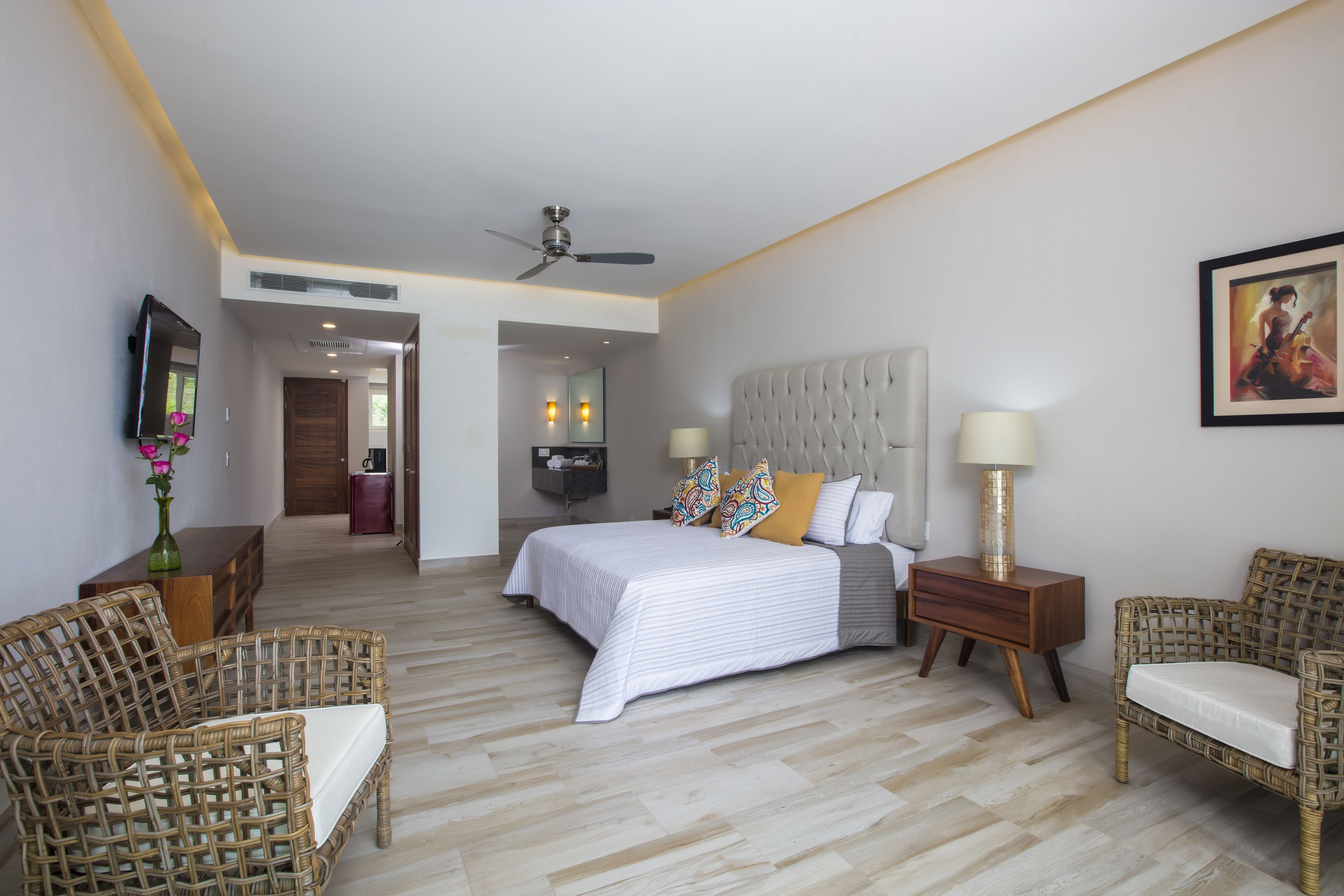 A modern hotel room with a large bed, two wicker chairs, a ceiling fan, and a TV on the wall.