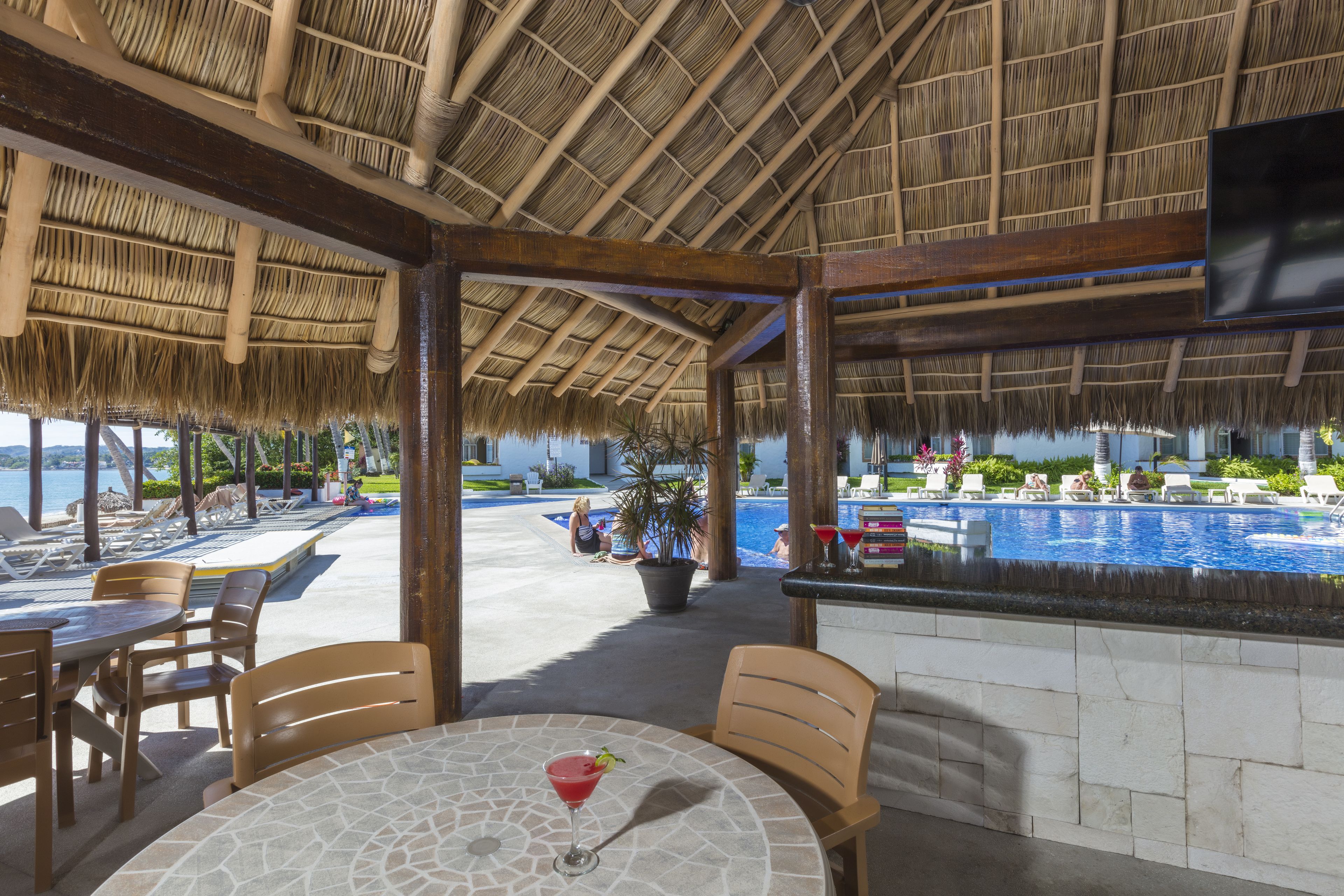 A covered outdoor seating area with a pool view, wooden furniture, and a stone table with a drink.