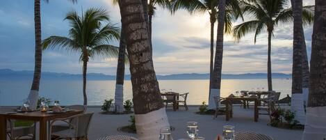 An outdoor dining area with tables and chairs, palm trees, and a view of the sea.