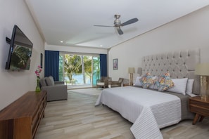 A spacious bedroom with a large bed, a ceiling fan, a TV mounted on the wall, and a view of palm trees through the window.