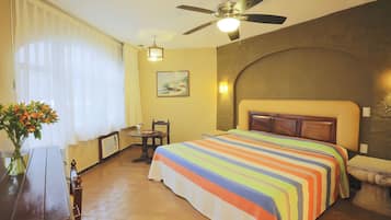 A hotel room with a bed, a desk, a chair, a vase with flowers, a ceiling fan, and a painting on the wall.