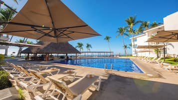 Un área de piscina con sillas de descanso y sombrillas, una estructura de techo de paja y palmeras.