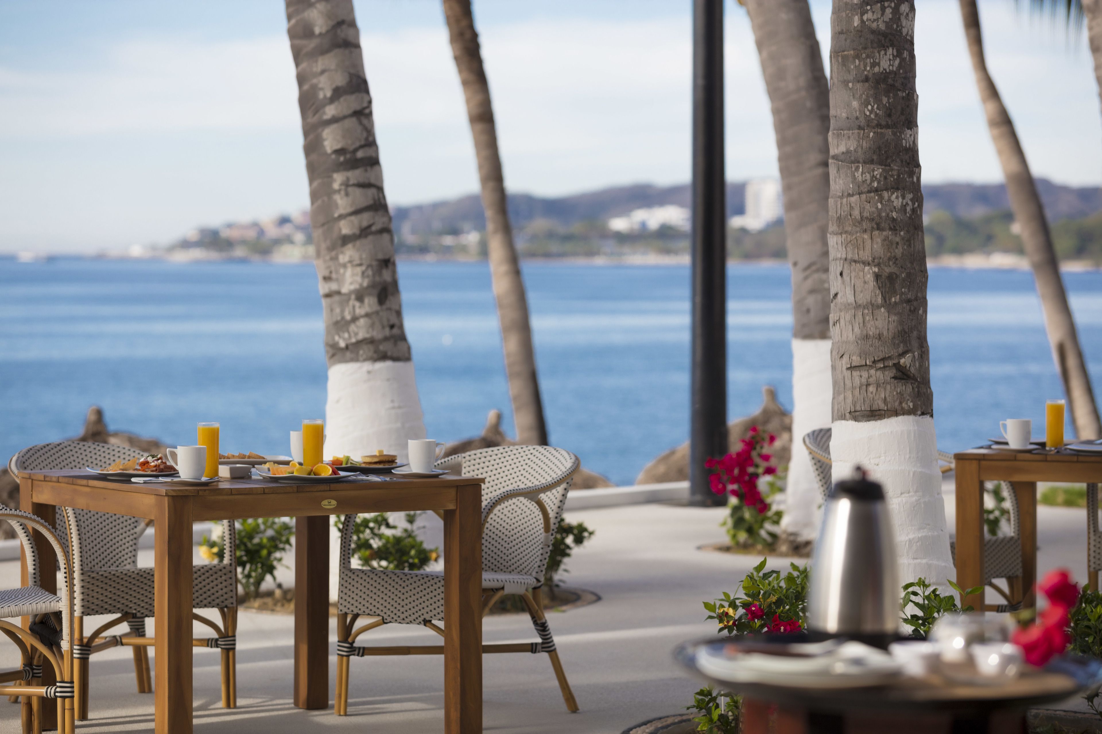 Un espacio para comer frente al mar, con sillas y mesas de mimbre, una cafetera y vistas al océano.