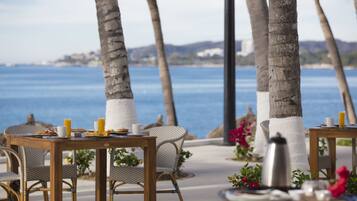 A seaside dining area with wicker chairs and tables, a coffee pot, and a view of the ocean.