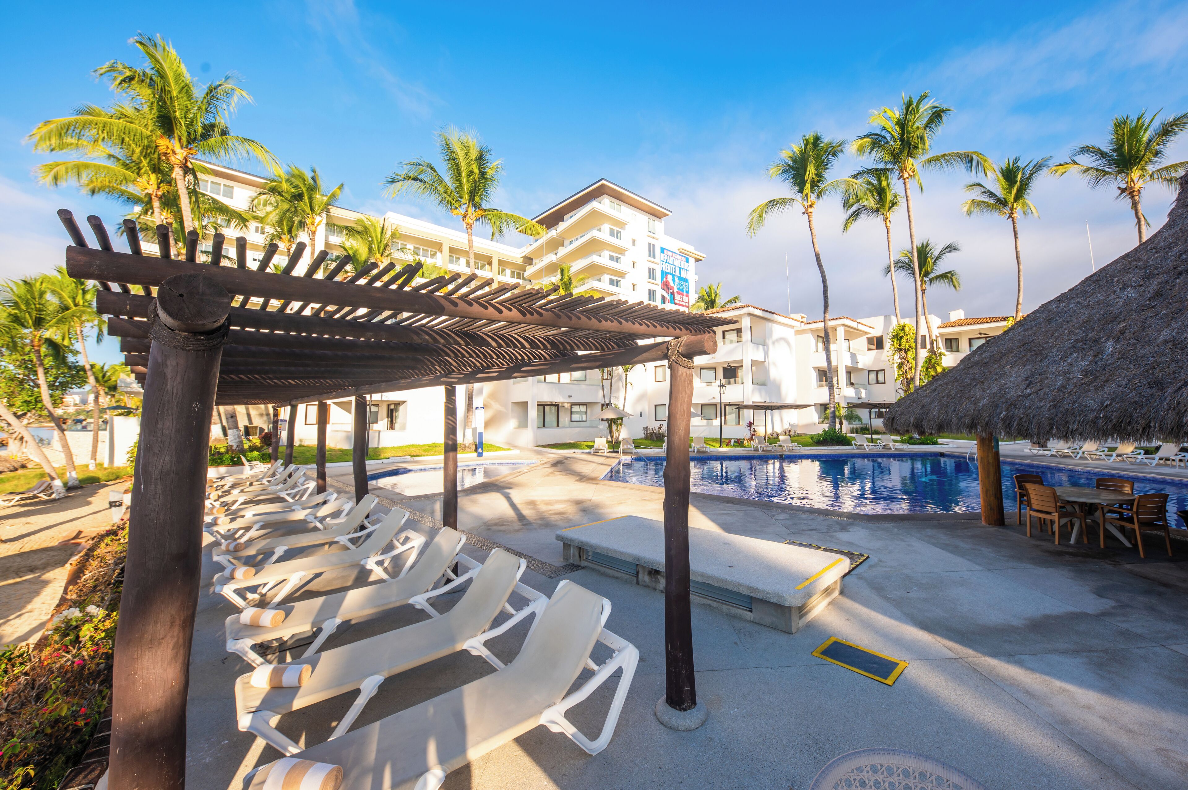A poolside area with lounge chairs, a pergola, a pool, and palm trees.