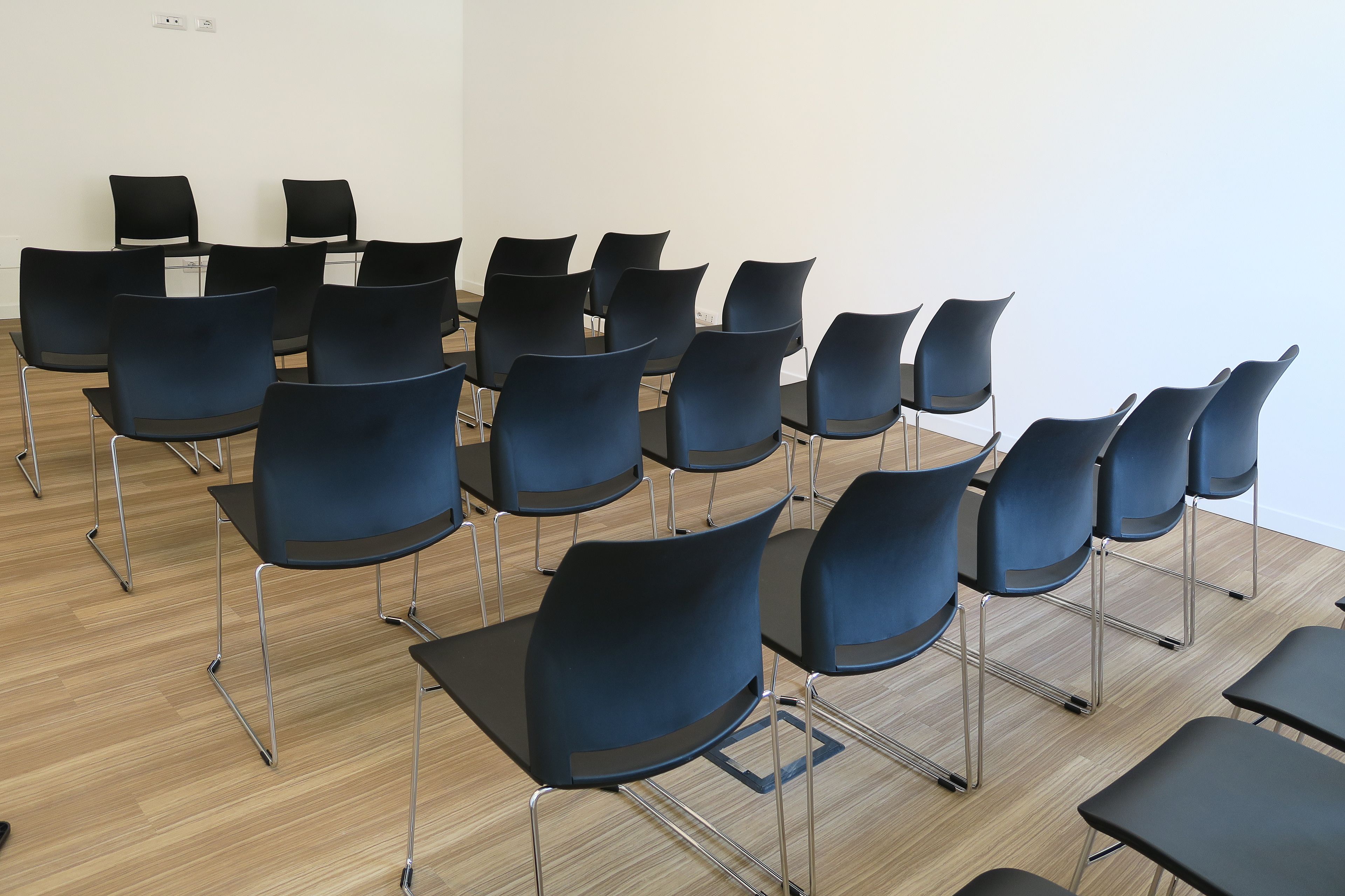 A room with rows of black chairs and tables, wooden flooring, and white walls.
