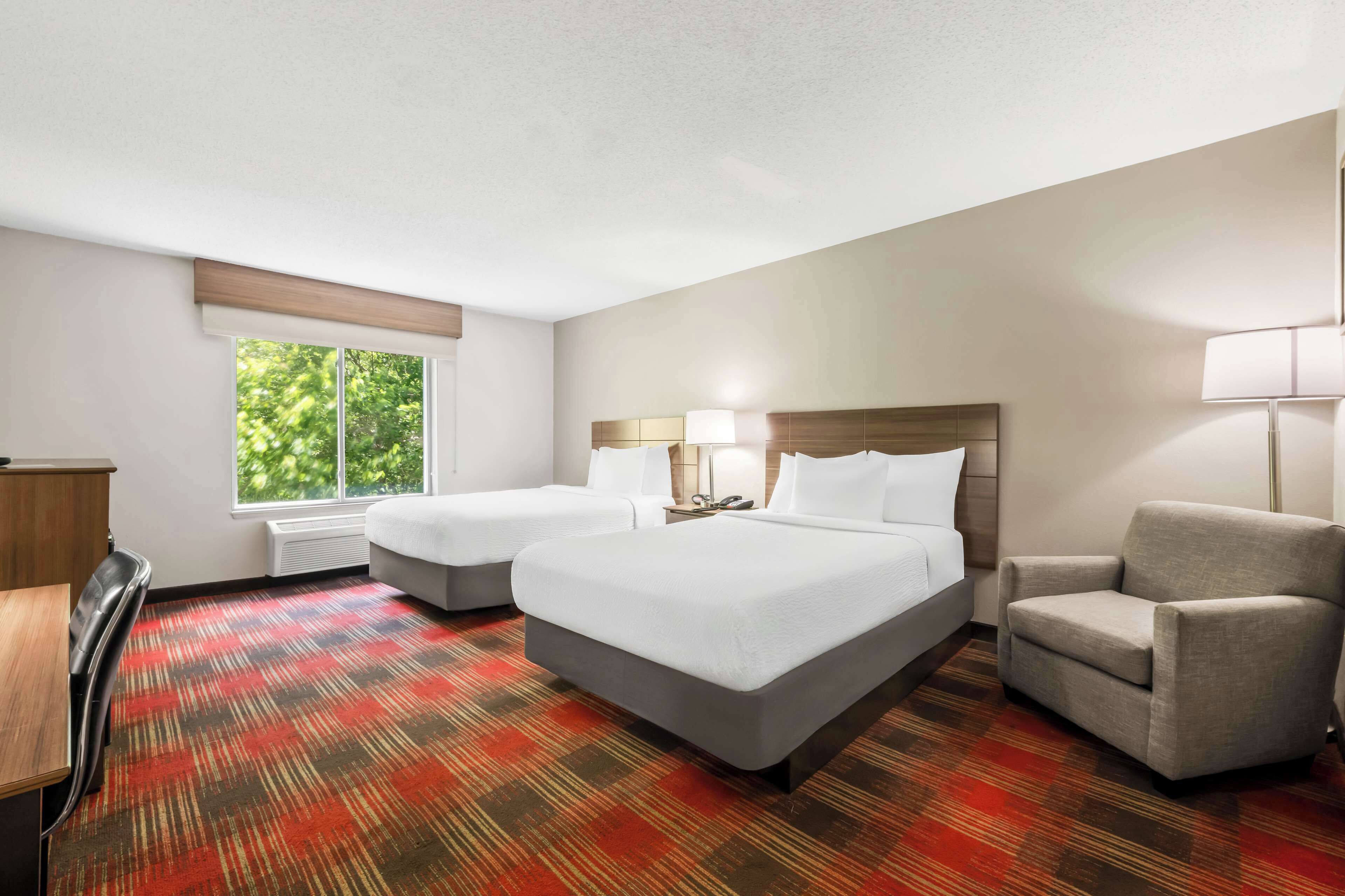 A hotel room with two beds, a desk, a chair, and a window with a view of trees.