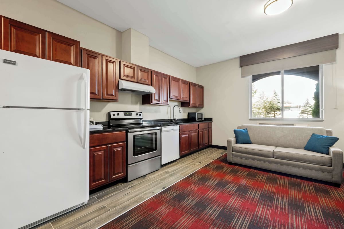 A kitchen with wooden cabinets, a white refrigerator, a stove, and a dishwasher. There is a grey sofa with blue cushions in the living area.