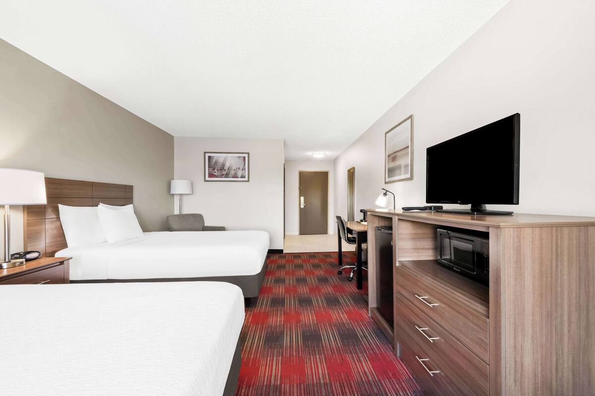 A hotel room with two beds, a desk with a flat-screen TV, a microwave, and a red carpeted floor.