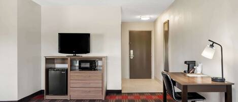 Habitación de hotel con kitchenette, televisor de pantalla plana, escritorio con lámpara y puerta que conduce a otra habitación.