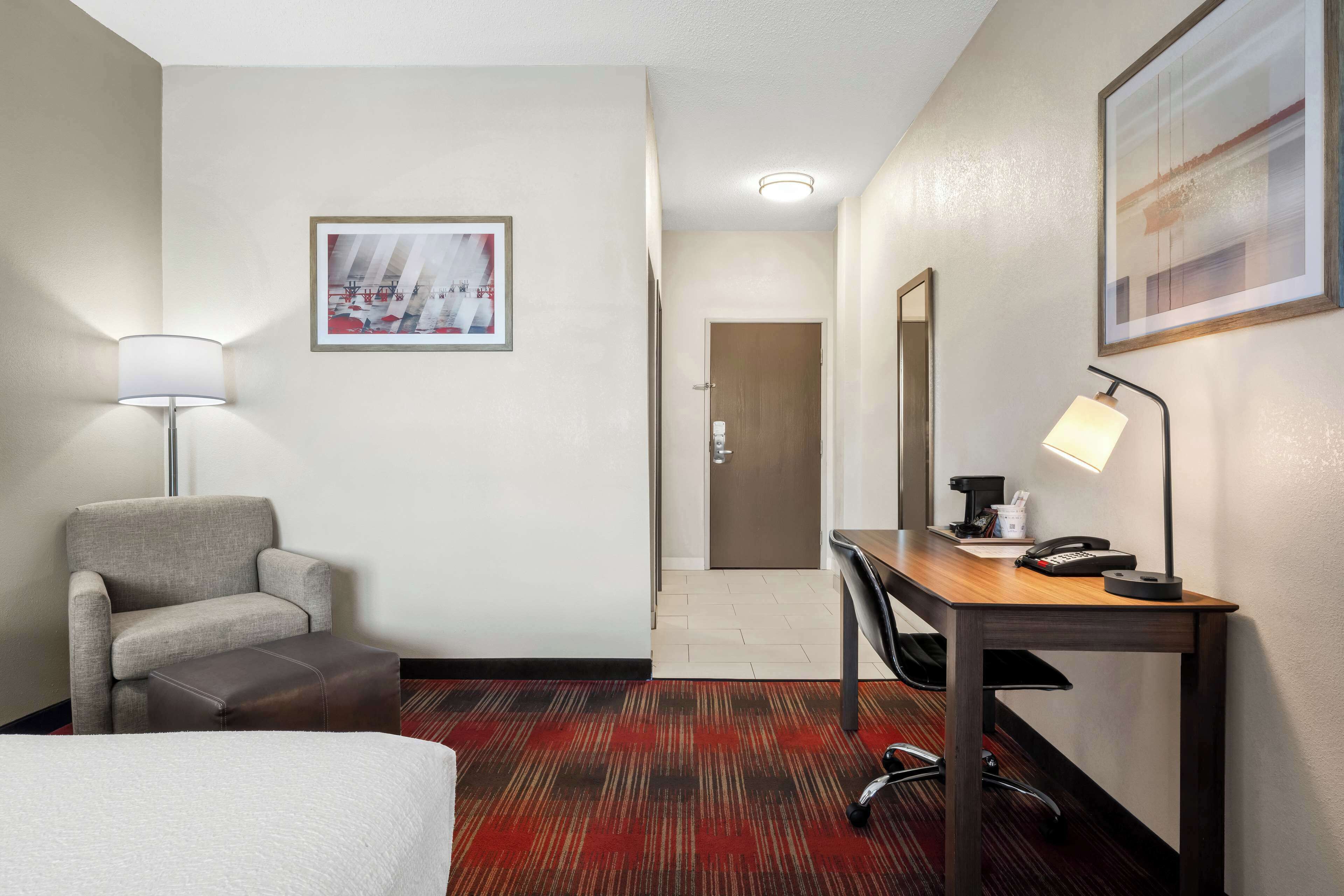 A hotel room with a bed, a desk, a chair, a lamp, and a door.