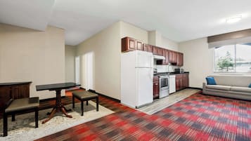 A modern kitchen with a refrigerator, stove, and wooden cabinets. There is a dining table with chairs and a sofa in the living area.