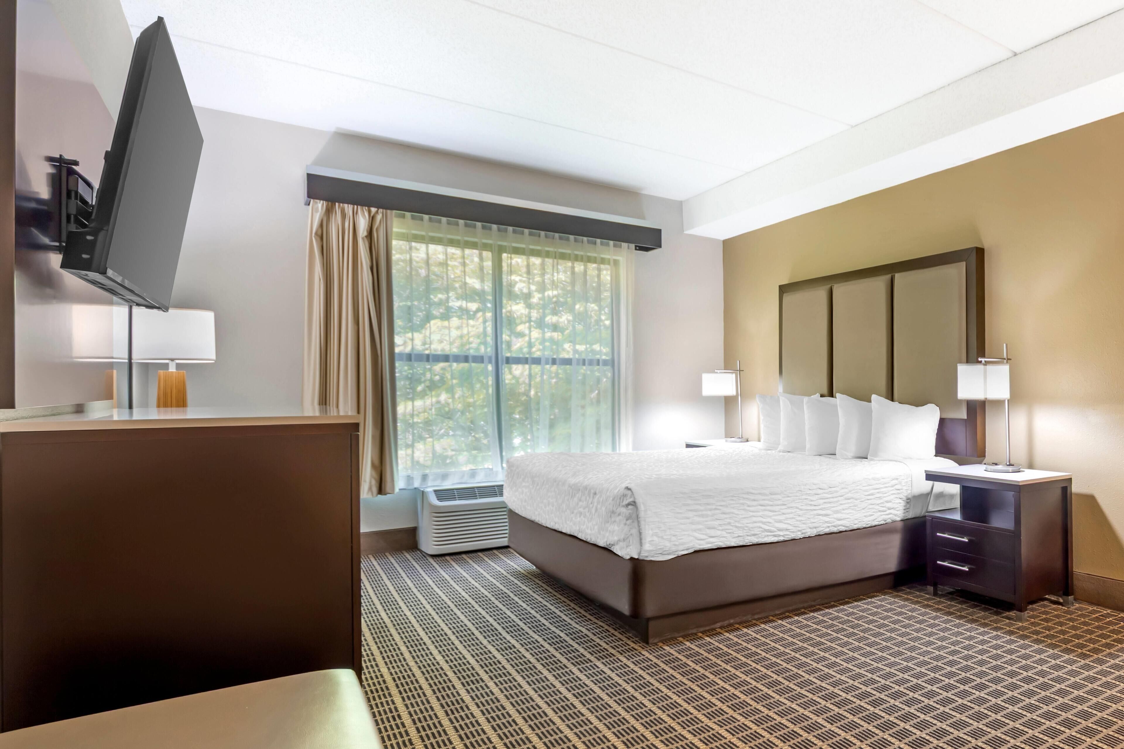 Una habitación de hotel con cama, lámparas de noche, televisión y un ventanal con cortinas.