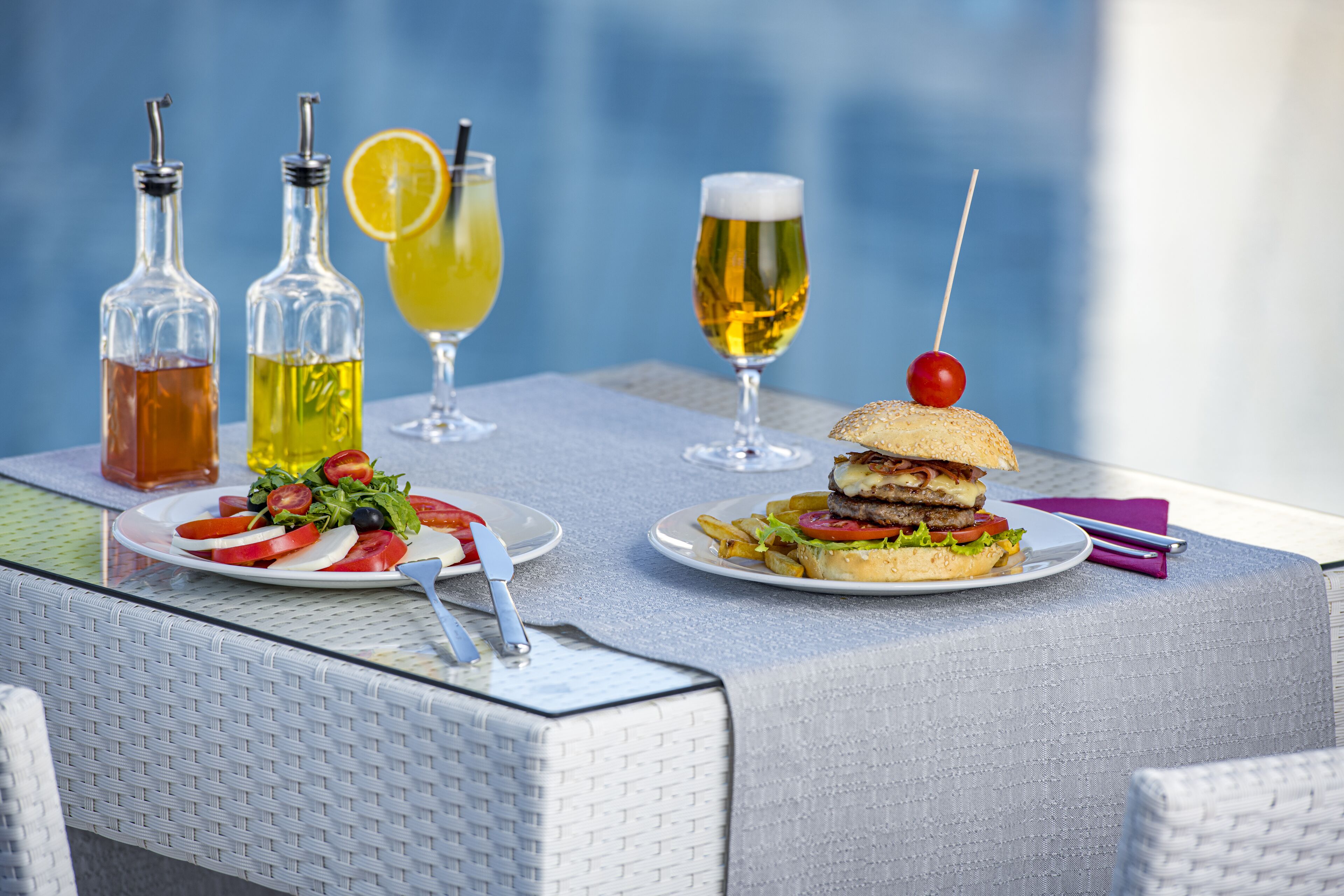Una mesa con ensalada, hamburguesa y bebidas, incluyendo cerveza y un cóctel con una rodaja de naranja.