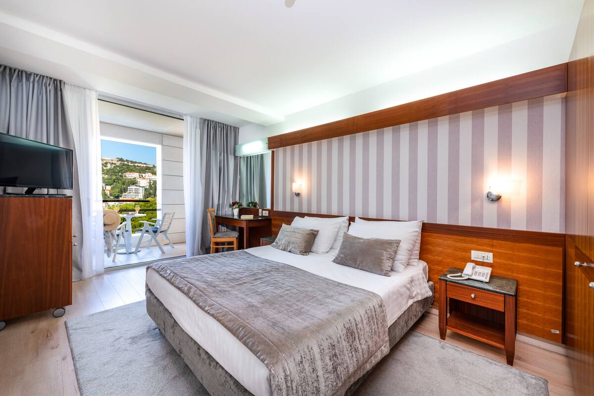 A modern hotel room with a large bed, a desk, a TV, and a balcony with a view of the city.