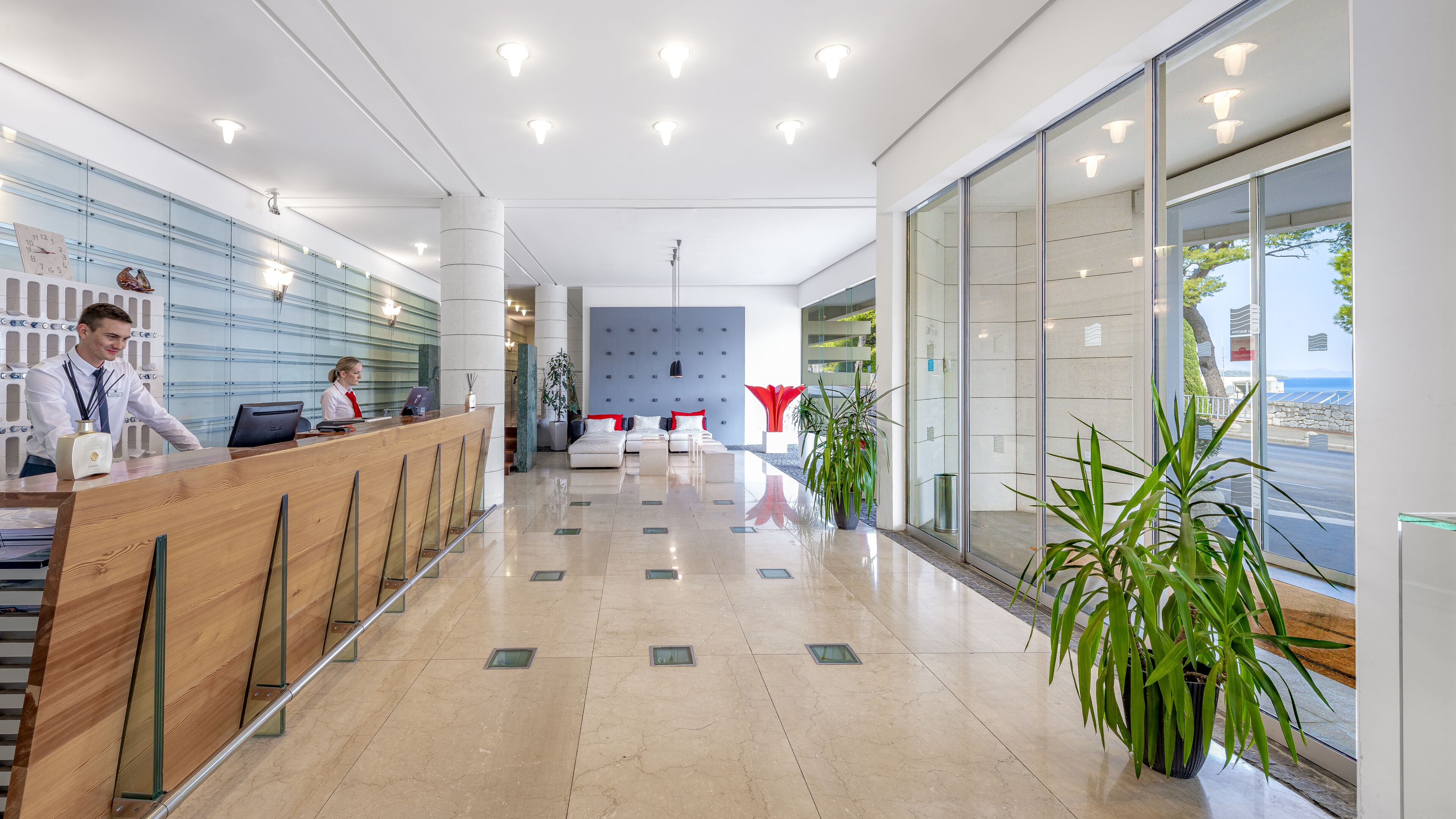A modern hotel lobby with a reception desk, seating area, and large windows offering a view of the sea.