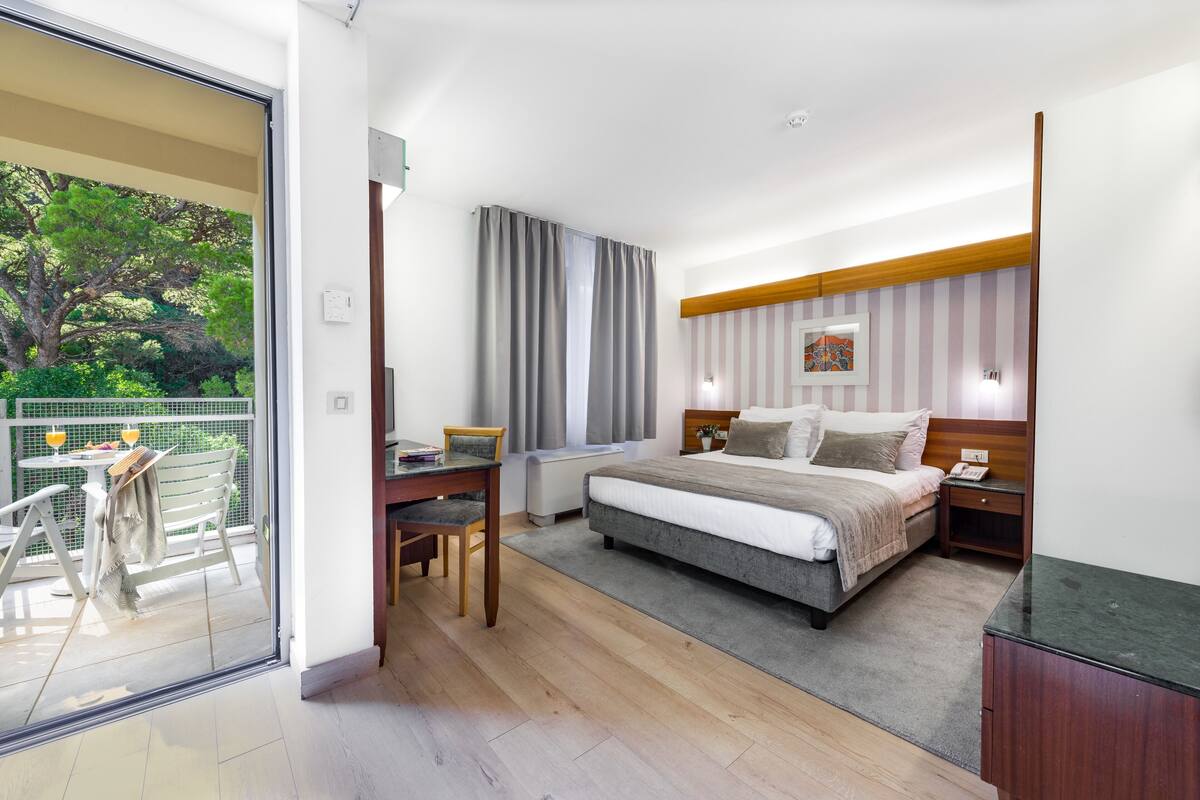 A modern hotel room with a large bed, a desk, and a balcony with a view of trees.