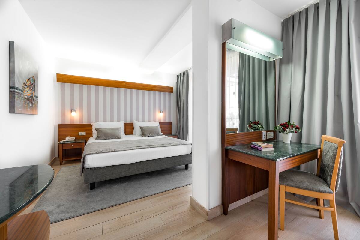 A modern hotel room with a bed, bedside table, desk, chair, and a large mirror.