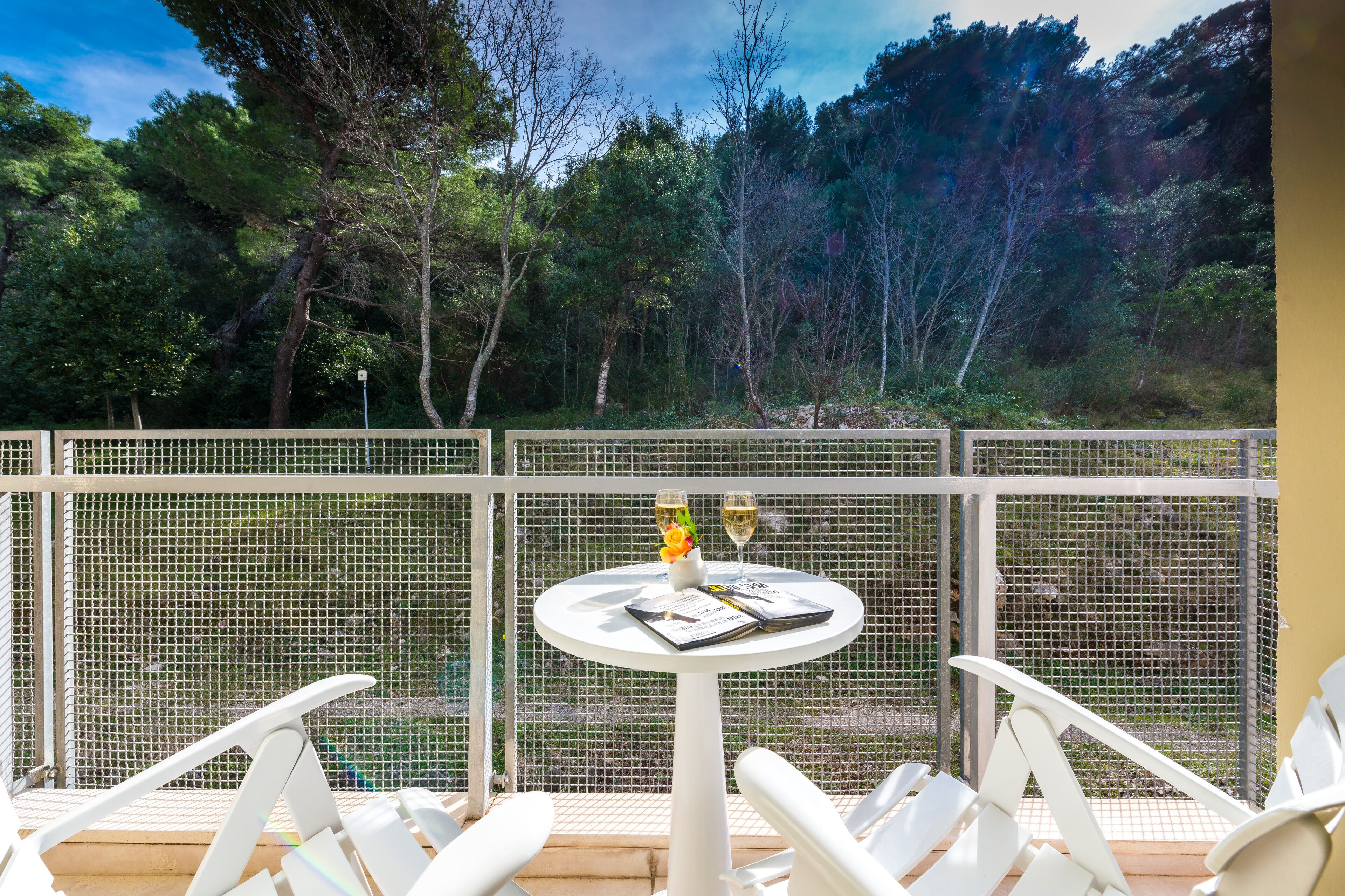 A balcony with white chairs and a table, a white vase with flowers, and two glasses of wine.