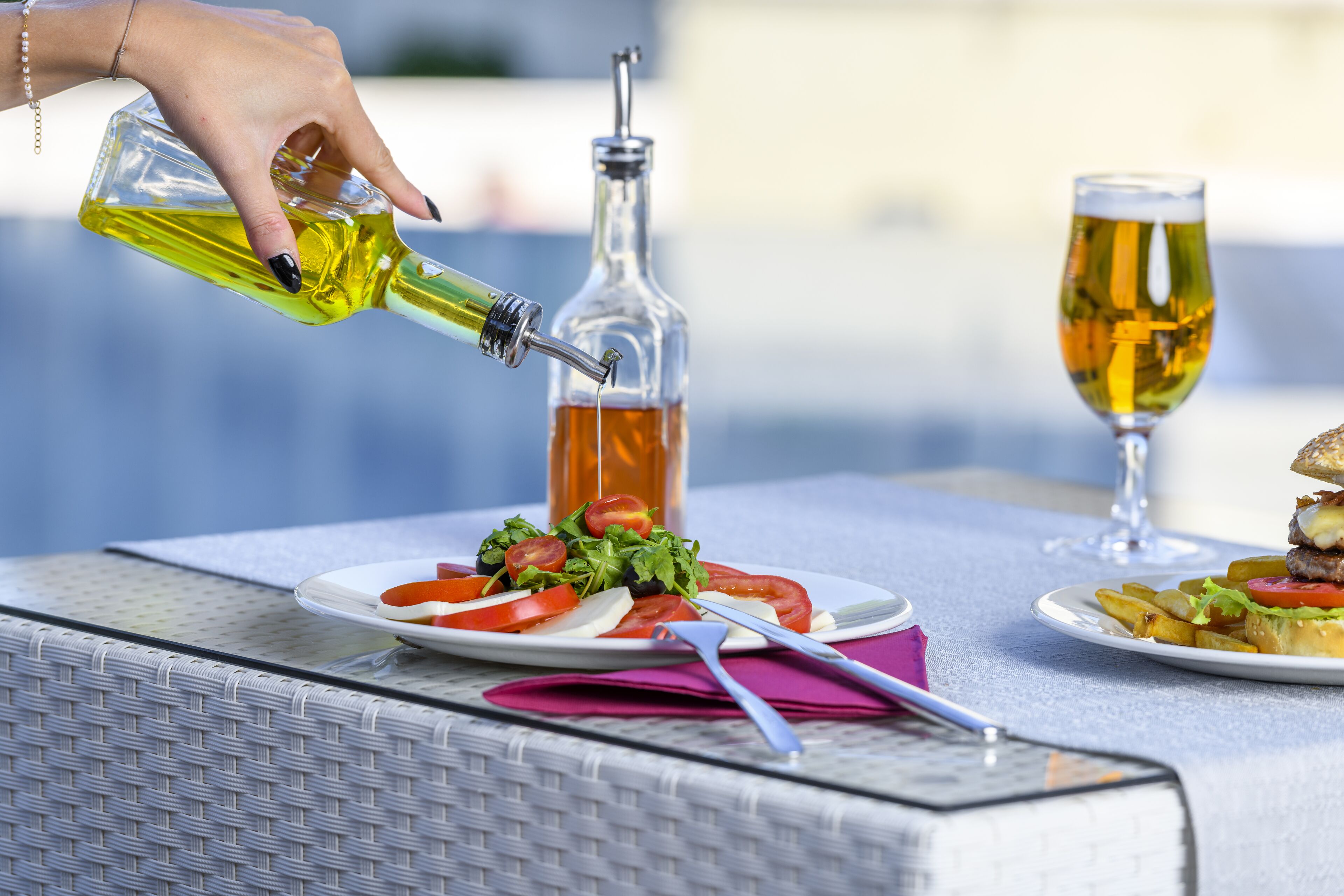 A table with a bottle of olive oil, a glass of beer, and plates of food including a burger and salad.