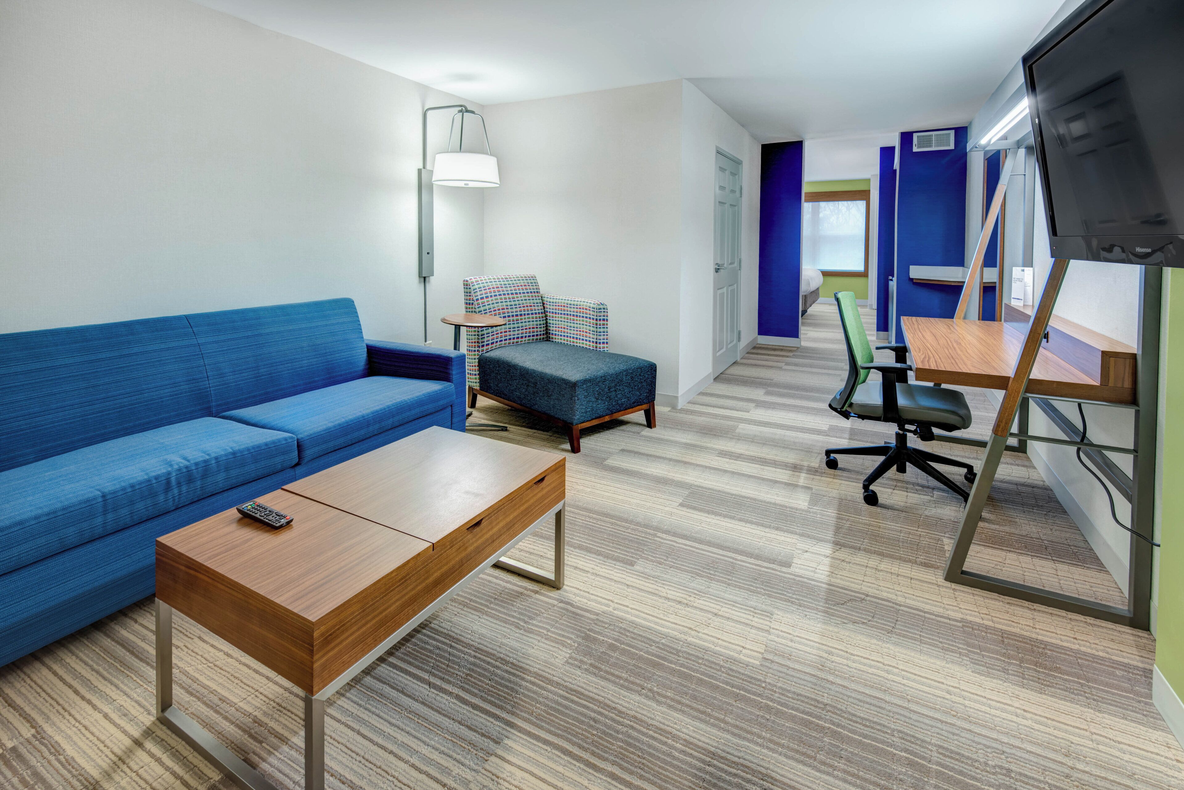 Moderni hotellihuone, jossa on sininen sohva, pieni pöytä, tuoli, työpöytä tietokoneella ja televisio.