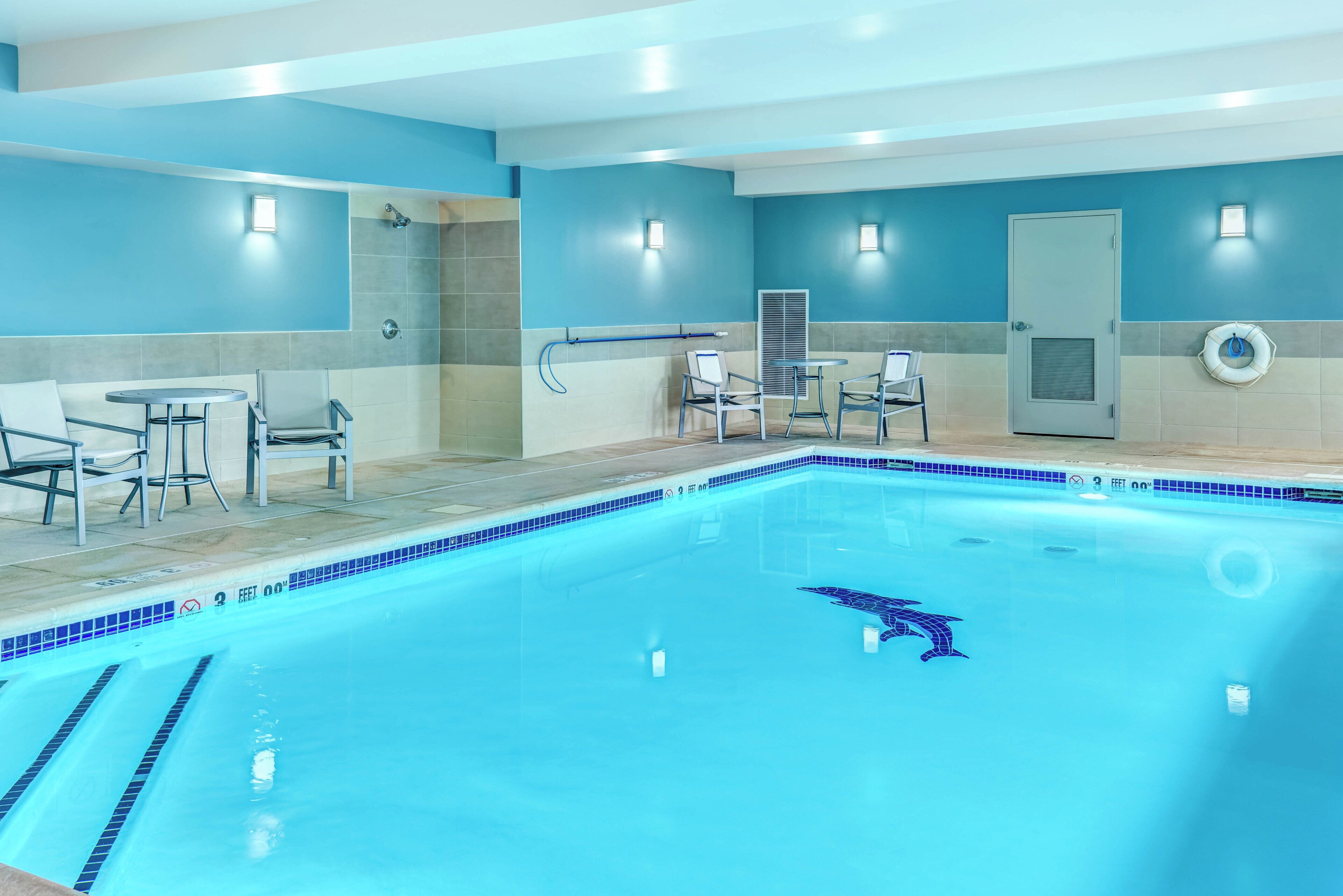 A swimming pool with a dolphin design, surrounded by chairs and a lifebuoy, with a door and a towel rack in the background.