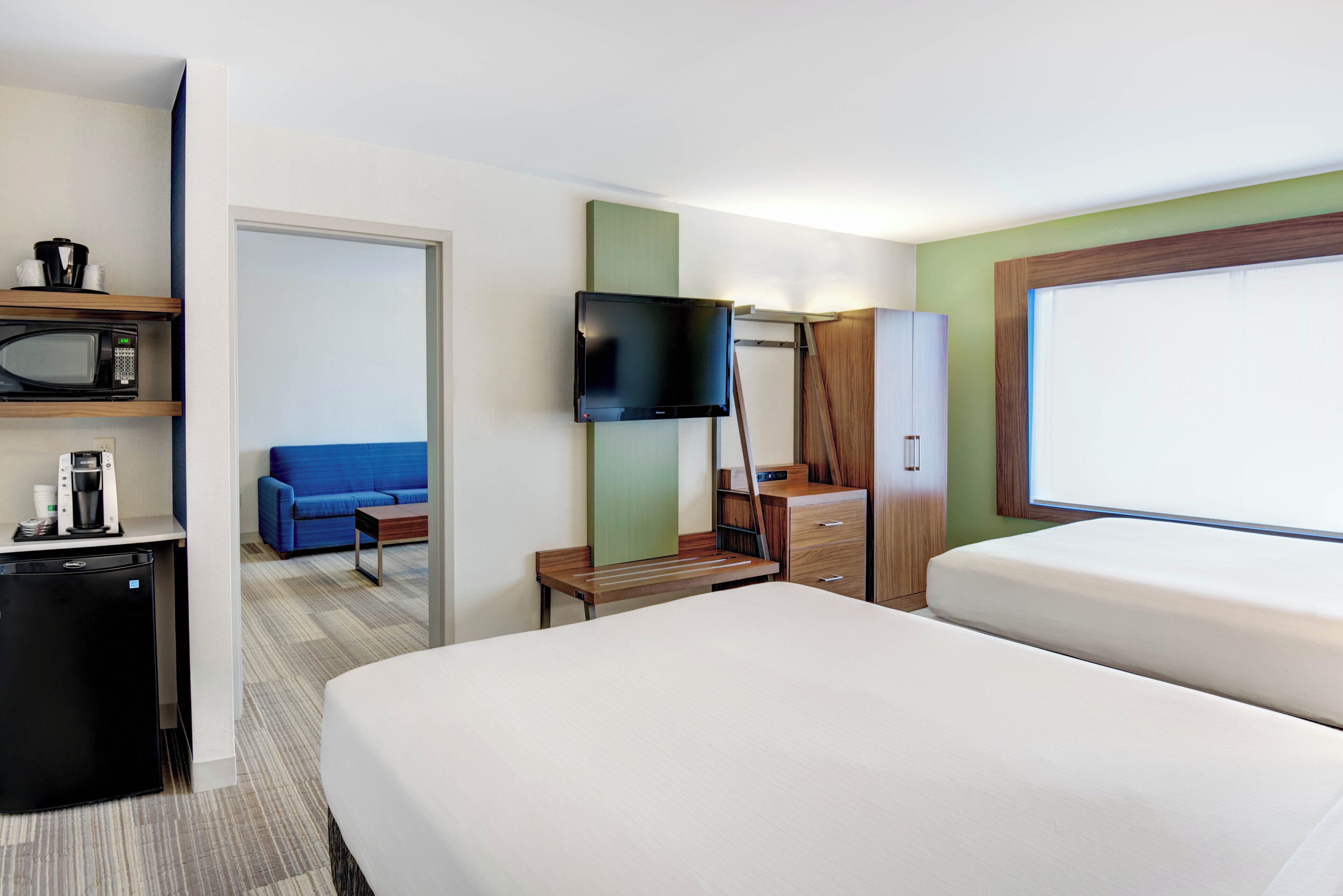 Una habitación de hotel con una cama, un televisor montado en la pared, una cafetera, un microondas y un sofá azul en la habitación contigua.