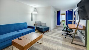 A modern room with a blue sofa, a wooden coffee table, a small blue armchair, a desk with a chair, and a TV mounted on the wall. - Holiday Inn Express Woodbridge by IHG (Avenel)