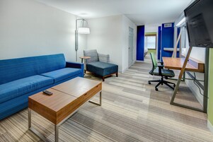 Una habitación moderna con un sofá azul, una mesa de centro de madera, un sillón azul pequeño, un escritorio con silla y un televisor montado en la pared.