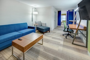 Una habitación moderna con un sofá azul, una mesa de centro de madera, un sillón azul pequeño, un escritorio con silla y un televisor montado en la pared.