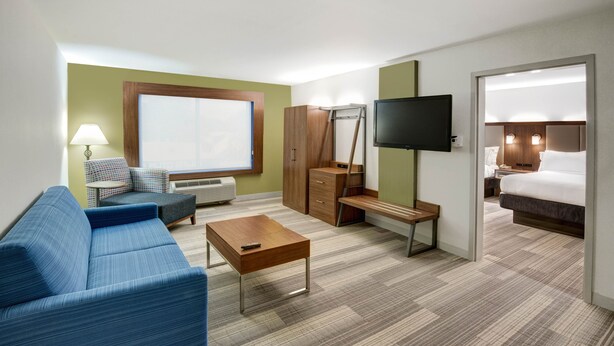 A hotel room with a blue sofa, a wooden coffee table, a desk, a wardrobe, a television, and a bed. - Holiday Inn Express Woodbridge by IHG (Avenel)